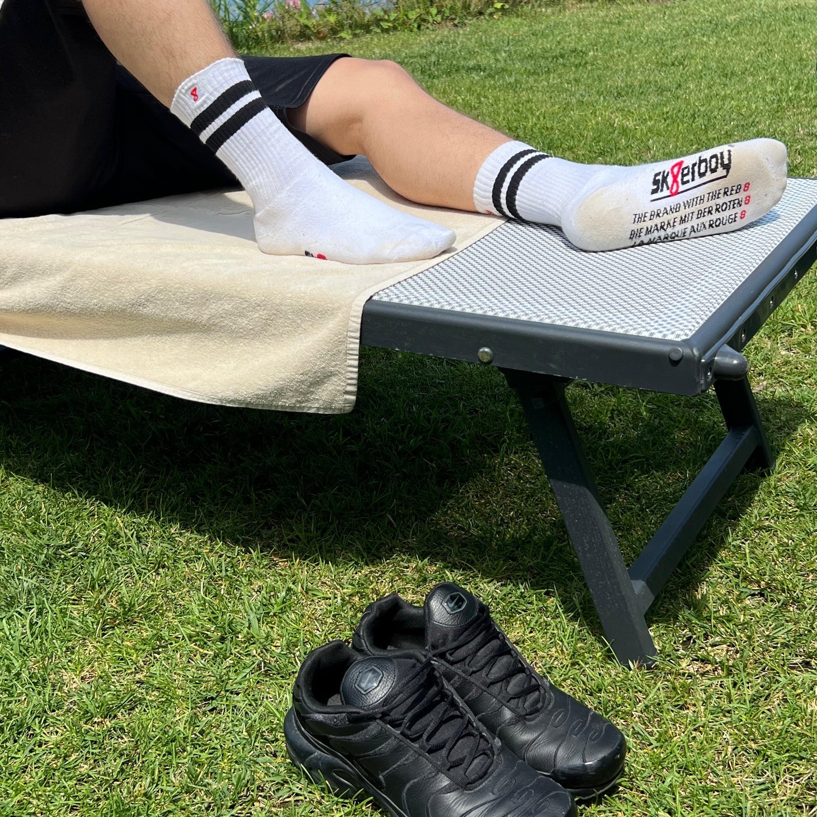 Person wearing socks with a brand logo, lying on a towel outdoors, with black shoes on the grass.
